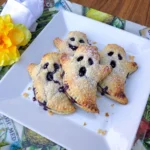 Four golden Blueberry Ghost Hand Pies with purple berry filling and ghost faces on a white plate